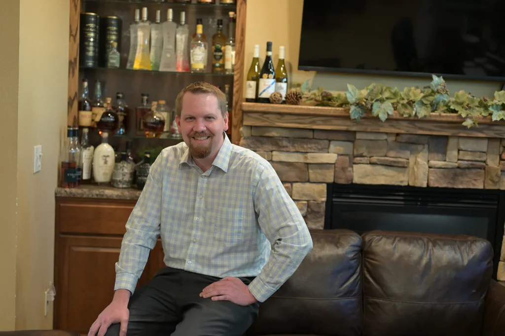 Kyle Lambert seated on leather couch in front of stone fire place with various bottles behind