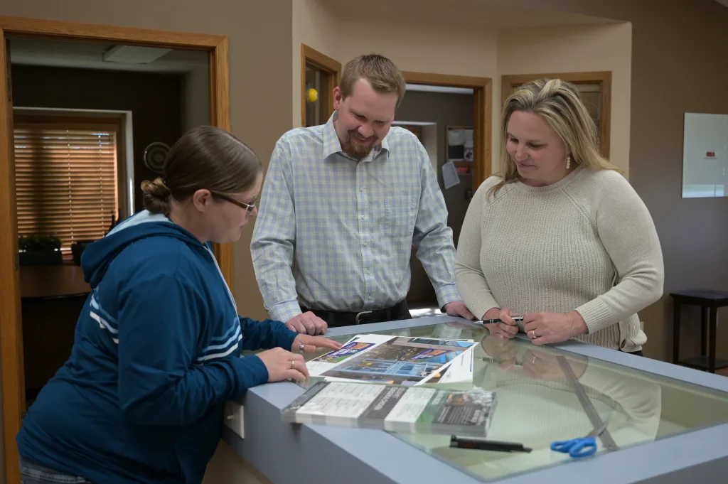 Three people stand around lighted table reviewing ad design assets