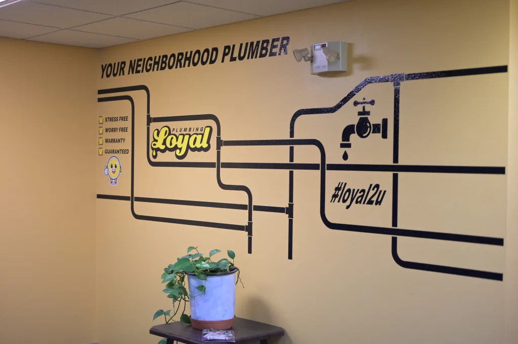 Brown wall with pipe design and words read Your Neighborhood Plumber, Loyal Plumbing logo, and #loyal2u