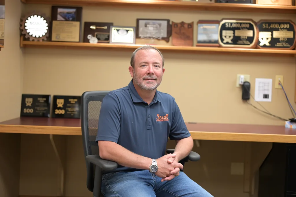 Andy Scull sits in office chair. Behind him are shelves with plaques and awards Scull Construction received.