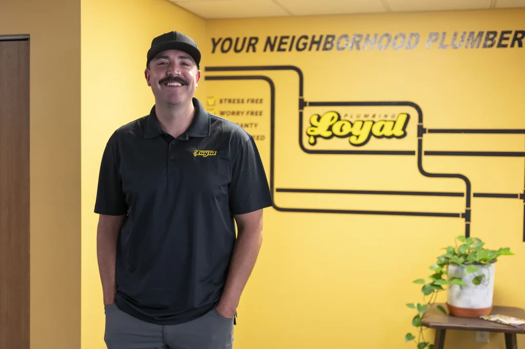 Mark Falcon stands in black polo shirt in front of yellow wall with the word Loyal behind him