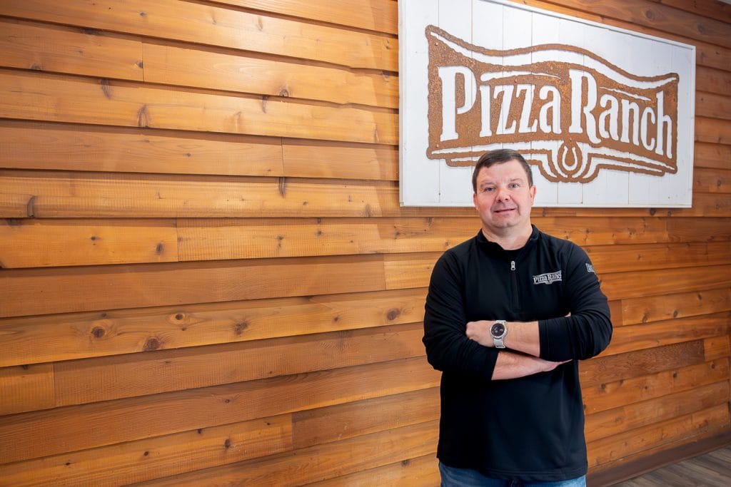 Owner Steven Cronin stands in front of wood wall with sign of pizza ranch logo