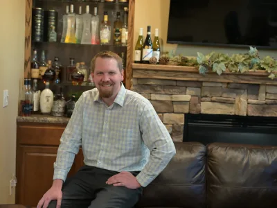 Kyle Lambert seated on leather couch in front of stone fire place with various bottles behind