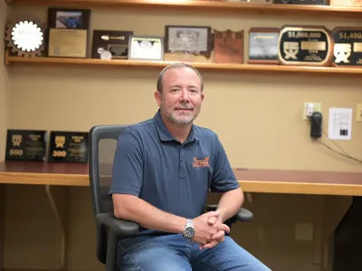 Andy Scull sits in office chair. Behind him are shelves with plaques and awards Scull Construction received.