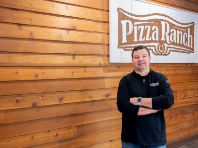 Owner Steven Cronin stands in front of wood wall with sign of pizza ranch logo